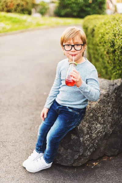 Venkovní portrét roztomilý chlapeček nosí brýle, světle modrý svetr, džíny a bílé košíky, drží nápoj s brčkem — Stock fotografie