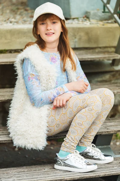 Outdoor fashion portrait of young preteen kid girl wearing white cap, faux fur gilet and silver mirror trainers — Stock Photo, Image