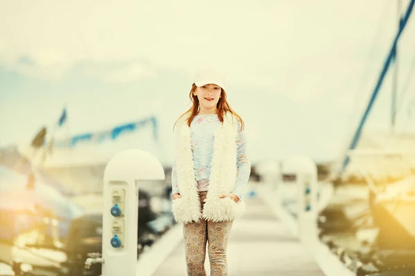 Outdoor fashion portrait of young preteen kid girl wearing white cap, faux fur gilet, playing in a port on a very sunny day, toned image — Stock Photo, Image