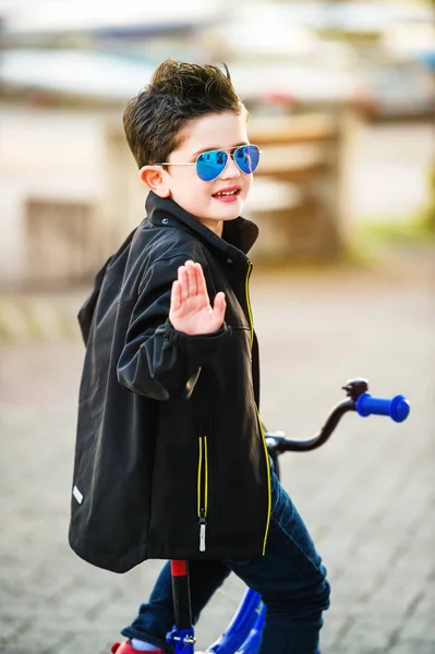 Małe dziecko chłopiec jazda na rowerze, patrząc wstecz na ramieniu, na sobie czarny płaszcz i okulary przeciwsłoneczne — Zdjęcie stockowe