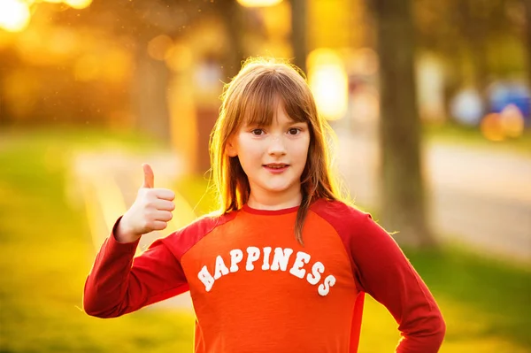 Menina bonito jogando ao ar livre em uma boa noite de verão, vestindo camiseta vermelha com sinal Felicidade, polegar grande para cima, luz do sol quente — Fotografia de Stock