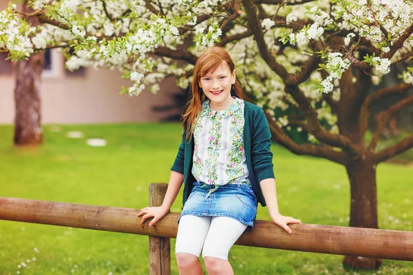 Frühlingsporträt von glücklichen jungen Mädchen, die auf dem Zaun sitzen, Jeansrock und grüne Jacke tragen — Stockfoto