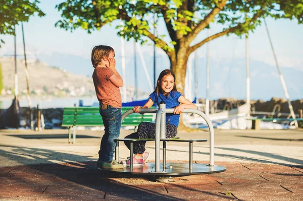 Két gyerek érzik magukat a játszótéren. Stílusos gyerek játszik a körhinta, a parkban nagyon süt a nap. Imádnivaló gyerekek meg együtt tölteni az időt — Stock Fotó