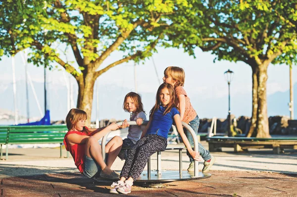 Grupo de cuatro niños divirtiéndose en el patio. Niños con estilo jugando en carrusel en el parque en un día muy soleado. Adorables niños amigos pasando tiempo juntos —  Fotos de Stock