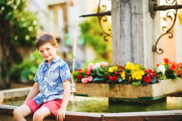 Sommer-Outdoor-Porträt eines entzückenden Jungen, der an einem sehr heißen, sonnigen Tag im Freien neben einem Brunnen ruht und ein blaues Hemd trägt — Stockfoto