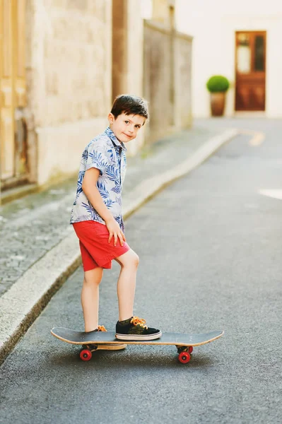 Funny stylish kid boy posing with skateboard outdoors, wearing blue print shirt and red shorts — Stock Photo, Image