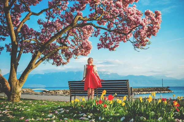 Ung blond kvinna du beundrar den fantastiska utsikten över Lake Geneva, Schweiz. Kvinna sitter på bänken under blossom magnoliaträd — Stockfoto