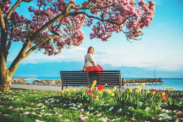 Fiatal szőke nő gyönyörködni a csodálatos kilátás nyílik a Genfi-tó, Svájc. Nő ül a padon, Virágzás magnólia fa alatt — Stock Fotó