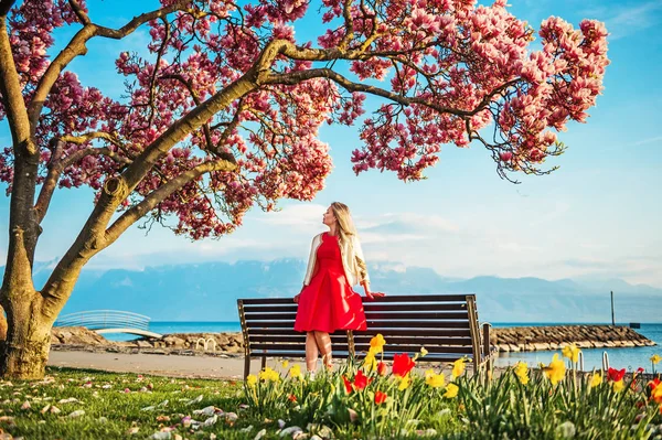 Młoda blond kobieta, podziwiając widok na Jezioro Genewskie, Szwajcaria. Kobieta siedzi na ławce pod kwitnąć magnolia drzewa — Zdjęcie stockowe