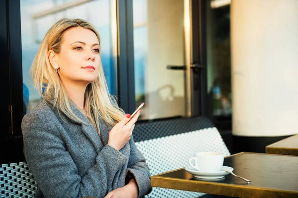 Beautiful blond woman resting in a coffee shop with cup of coffee on the table, holding smart phone in hands