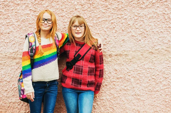 Grupo de dos niñas adorables posando al aire libre contra la pared rosa, con gafas, mochilas escolares y pulóveres de colores brillantes, de vuelta al concepto de la escuela —  Fotos de Stock