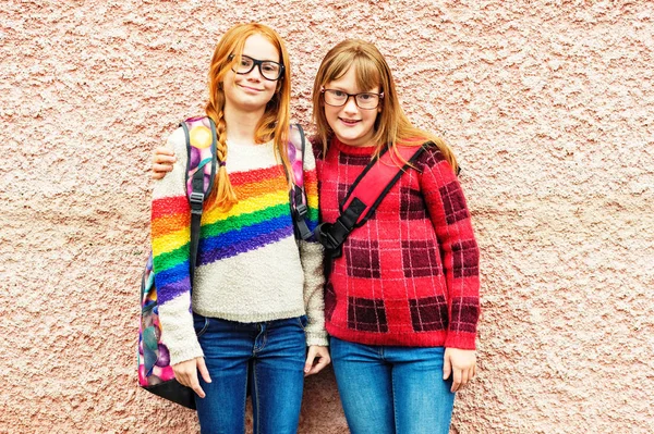 Grupo de dos niñas adorables posando al aire libre contra la pared rosa, con gafas, mochilas escolares y pulóveres de colores brillantes, de vuelta al concepto de la escuela —  Fotos de Stock