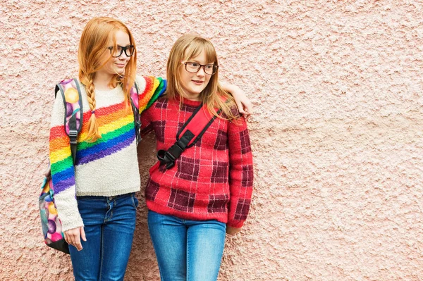 Grupo de dos niñas adorables posando al aire libre contra la pared rosa, con gafas, mochilas escolares y pulóveres de colores brillantes, de vuelta al concepto de la escuela —  Fotos de Stock