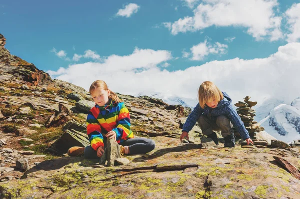 Niedliche kleine Kinder, die sich auf dem Gornergrat-Gletscher ausruhen, zwei kleine Kinder, die zusammen in den Bergen spielen, kleiner Junge und seine grosse Schwester, die in bunten Klamotten in den Schweizer Alpen wandern — Stockfoto