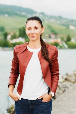 Kahverengi deri ceket giyen göl kenarında oturan güzel esmer kadın