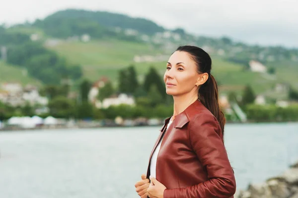 Kahverengi deri ceket giyen göl kenarında oturan güzel esmer kadın — Stok fotoğraf