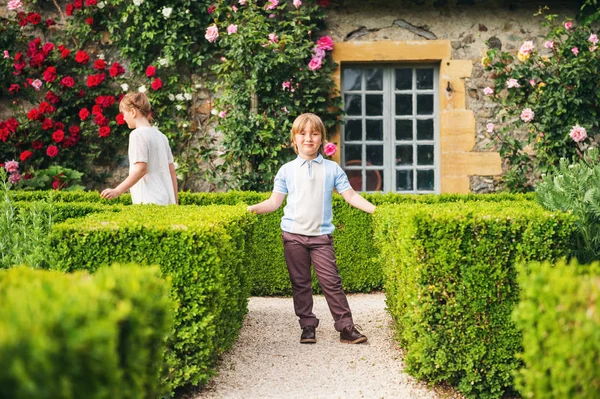 Två barn, liten pojke och flicka, poserar i vacker klassisk engelsk figurklippning trädgård, retro stil kläder. Bror och syster spelar tillsammans i fantastisk sommar park mellan blommande rosor — Stockfoto