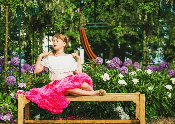 Menina engraçada descansando no banco em um belo jardim de flores em um bom dia de verão, vestindo camisa branca e saia de tutu rosa brilhante — Fotografia de Stock