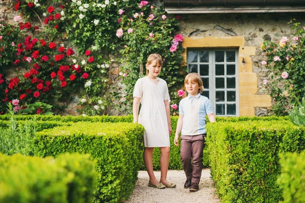 Two kids, little boy and girl, posing in beautiful classical English topiary garden, wearing retro style clothing. Brother and sister playing together in amazing summer park between blooming roses Stock Picture