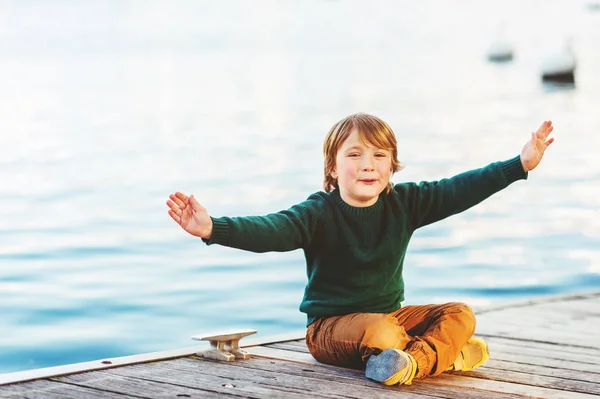 Retrato ao ar livre de adorável menino de 5-6 anos descansando junto ao lago, vestindo pulôver verde e calças amarelas, braços abertos — Fotografia de Stock