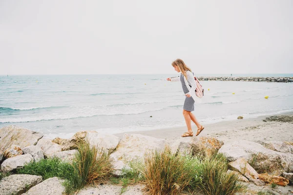 Маленькая девочка наслаждается летним отдыхом у моря, одетая в полосатое морское платье и рюкзак. Изображение сделано в Сент-Мари-де-ла-Мер, столице Камарга, на юге Франции — стоковое фото