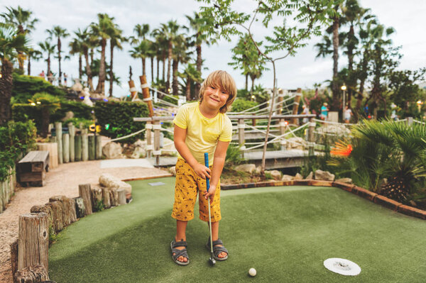 Забавный мальчик играет в мини-гольф, ребенок наслаждается летними каникулами
