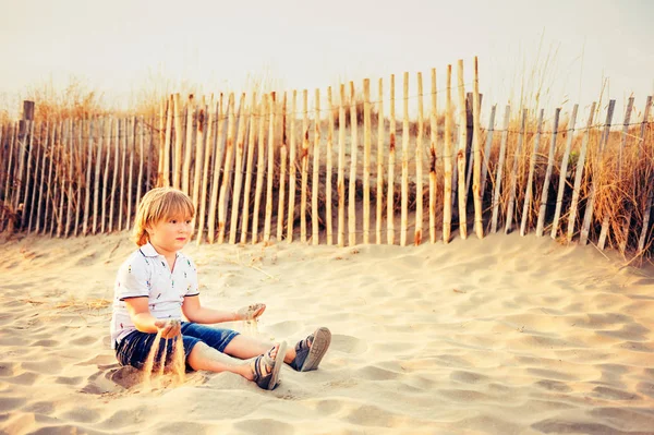 Grappige jongen jongen rusten op zand strand aan zee bij zonsondergang. Jong kind genieten van zomer vakantie op ten zuiden van Frankrijk, dragen polo, denim shorts en sandalen schoenen — Stockfoto