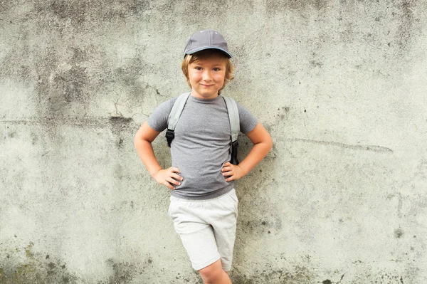 Πορτραίτου του αστεία μικρό αγόρι που στέκεται δίπλα σε γκρίζο τοίχο, φορώντας το καπάκι και σακίδιο — Φωτογραφία Αρχείου