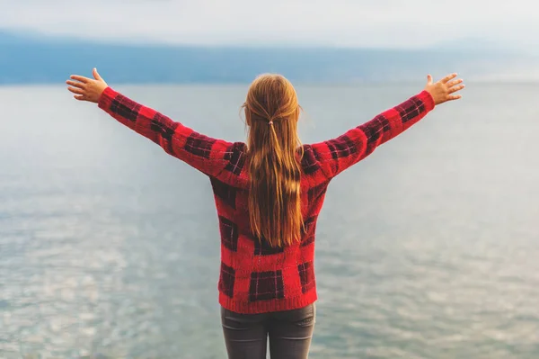 Kid girl enjoying nice autumn day ba the lake; wearing warm red pullover, arms up wide open, back view. — Stock Photo, Image