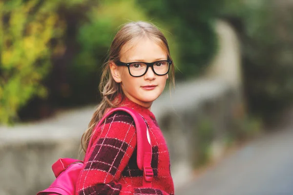 Outdoor portrait of a cute little 9 year old girl, wearing red pullover, glasses and pink backpack — Stock Photo, Image