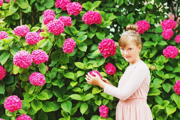 Outdoor portrait of pretty little girl playing with beautiful hydrangea flowers in summer garden, wearing ballerina style dress, hair bun and diamond tiara — Stock Photo, Image