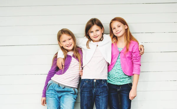 Denim kot pantolon ve renkli ceket giyen 3 küçük kız açık havada beyaz ahşap arka plan karşı ayakta grubu — Stok fotoğraf