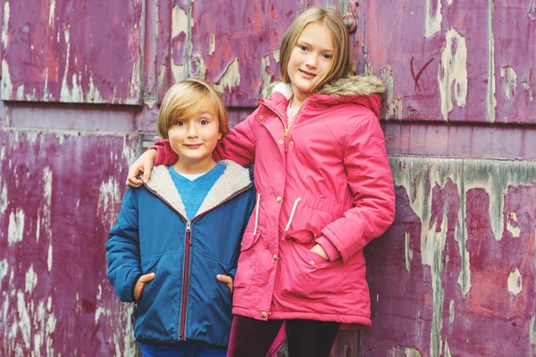 Портрет двух очаровательных детей на улице, одетых в теплые пальто, стоящие рядом со старым фиолетовым фоном — стоковое фото