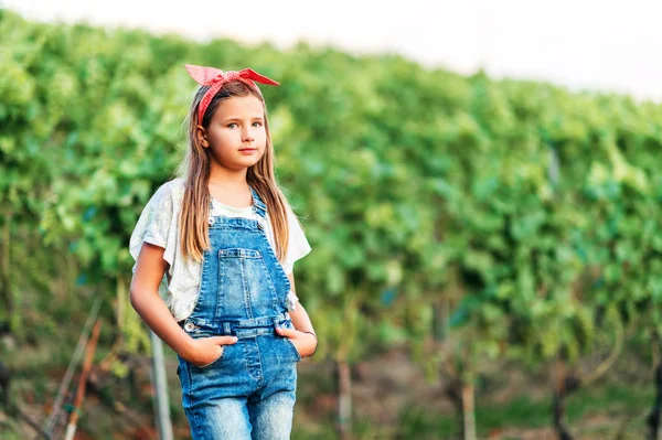 Outdoor portrait of pretty little girl wearing denim overalls and red vintage headband, hands in pockets — Stock Photo, Image