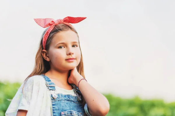 Outdoor portrait of pretty little girl wearing denim overalls and red vintage headband, hands in pockets — Stock Photo, Image
