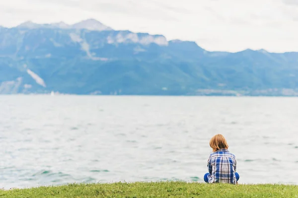 Utomhus porträtt av lilla kid boy spelar vid sjön på en molnig dag, sitter på gräset, klädd i blå tröja. Bakifrån. Bild tagen vid Genèvesjön, Lausanne, Schweiz — Stockfoto