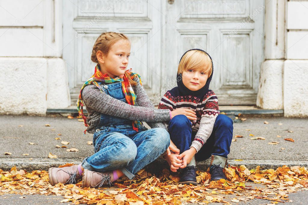Autumn portrait of 2 adorable kids in a city, wearing warm pullovers and denim jeans