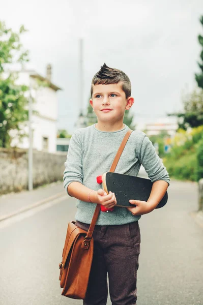 Outdoor portrait of funny little schoolboy wearing brown leather bag over shoulder, holding skateboard. Back to school concept. Film look toned image — Stock Photo, Image