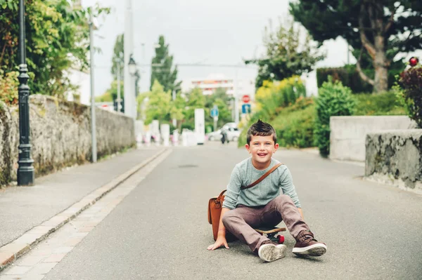 Outdoor portrait of funny little schoolboy wearing brown leather bag over shoulder, riding skateboard. Back to school concept. Film look toned image — Stock Photo, Image