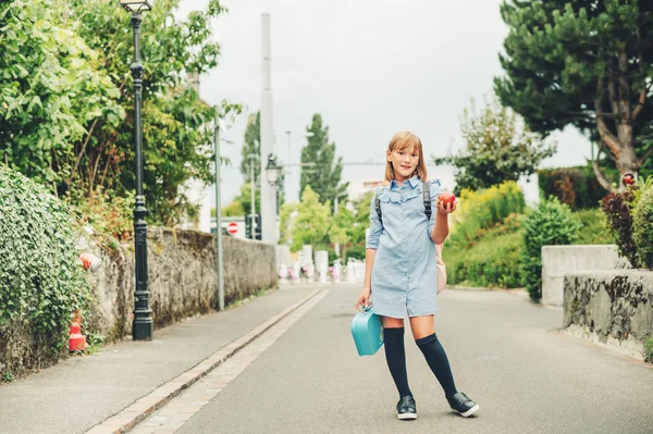 Retrato al aire libre de la colegiala divertida, con vestido formal y mochila, sosteniendo lonchera y manzana roja. Volver al concepto de la escuela, imagen de aspecto de película tonificada — Foto de Stock