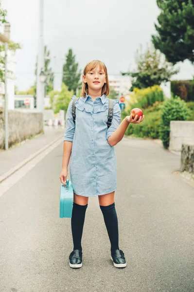Outdoor portrait of funny little schoolgirl, wearing formal dress and backpack, holding lunch box and red apple. Back to school concept, film look toned image — Stock Photo, Image