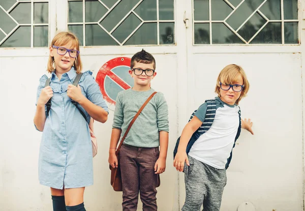 Group of three funny kids wearing backpacks walking back to school. Girl and boys wearing eyeglasses posing outdoors — Stock Photo, Image