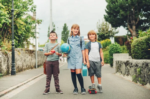 Un gruppo di tre ragazzi divertenti che indossano zaini mentre tornano a scuola. Ragazza e ragazzi che amano le attività scolastiche. Globo, cestino pranzo, mela rossa e accessori skateboard . — Foto Stock