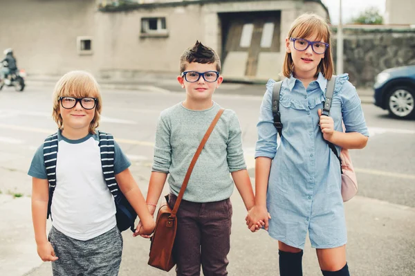 Group of three funny kids wearing backpacks walking back to school. Girl and boys wearing eyeglasses posing outdoors next to road — Stock Photo, Image