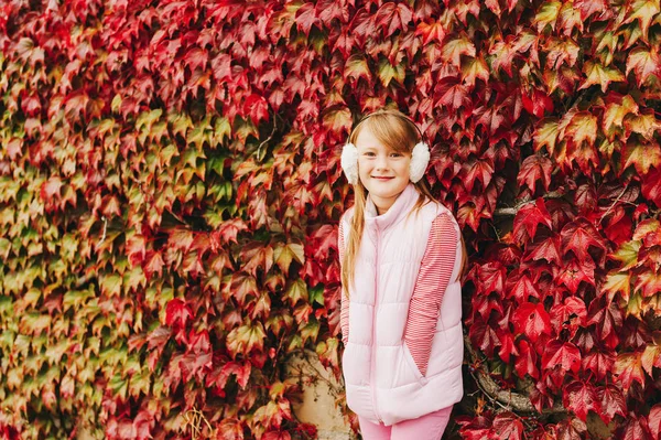 Ritratto all'aperto di bella bambina in posa contro il muro rosso ricoperto di foglie di edera rossa, indossando gilet rosa caldo e paraorecchie — Foto Stock