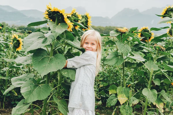 Adorable little 3-4 year old kid girl playing with giant sunflowers in a field. Happy childhood in a countryside. Autumn fashion for children — Stock Photo, Image