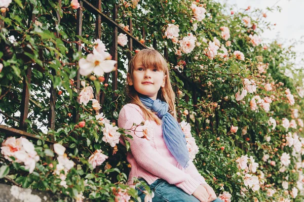 Ritratto all'aperto di una graziosa bambina di 7 anni che indossa un pullover rosa e una sciarpa blu, in posa tra morbide rose chiare. Moda autunno per bambini — Foto Stock