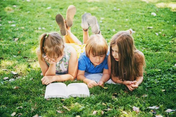 Gruppo di tre bambini sdraiati sull'erba verde e che leggono libri di storie insieme. Istruzione per i bambini — Foto Stock