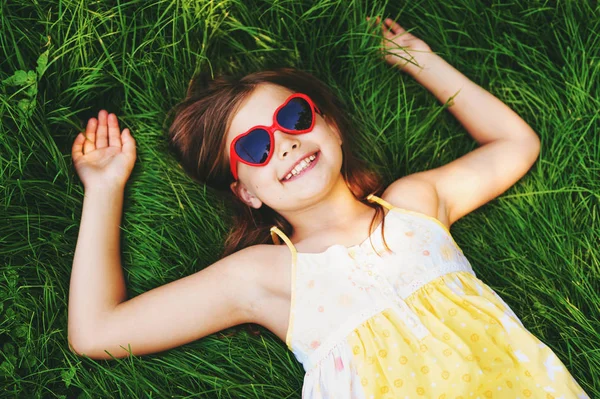 Outdoor portrait of pretty little girl wearing heart shaped sunglasses, lying on fresh green grass. Party for children, summer fun, happy childhood. Top view — Stock Photo, Image