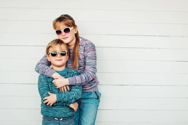 Groep van twee grappige kinderen samen buiten spelen, kleine jongen en meisje poseren tegen witte houten achtergrond, broer en zus dragen bijpassende truien, mode voor kinderen, breigoed. — Stockfoto